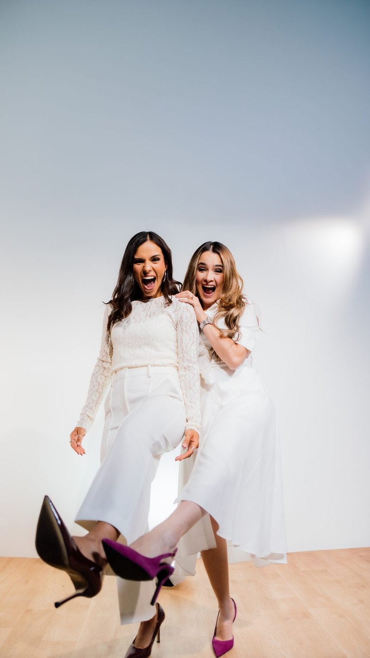 Dos mujeres vestidas de blanco, de cuerpo entero y riendo en un estudio de fondo claro.